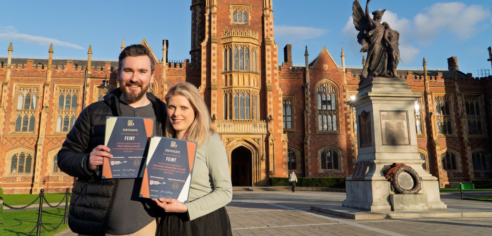 a smiling bearded man in a dark, short coat stands beside a smiling blonde woman, both holding purple certificates and standing in front of an old red brick building